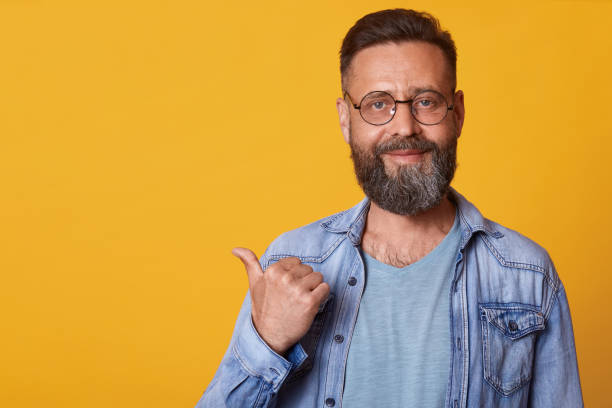 istock Happy optimistic handsome middle aged male with beard pointing aside with thumb and looking at camera, smiling man in denim jacket and gray t shirt. Copy space for promotin text or advertisment. 1153686326