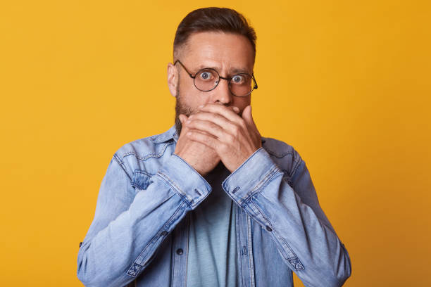 istock Shocked scared middle aged surprised face covering mouth with both hands, horrified stunned looking at camera isolated on yellow studio background, startled guy wearing denim kjacket and gray jacket. 1153686357