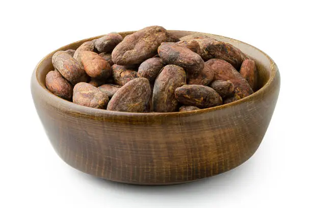 Roasted unpeeled cocoa beans in a brown wooden bowl isolated on white.