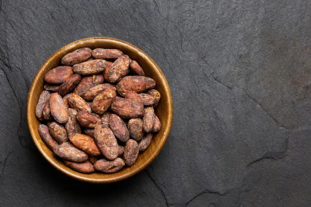 Roasted unpeeled cocoa beans in a brown wooden bowl isolated on black slate from above. Space for text.