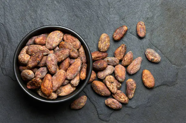 Roasted unpeeled cocoa beans in a black ceramic bowl next to unpeeled cocoa beans isolated on black slate from above.