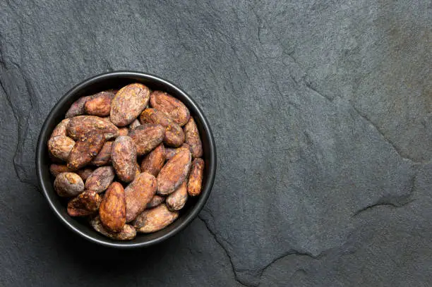 Roasted unpeeled cocoa beans in a black ceramic bowl isolated on black slate from above. Space for text.