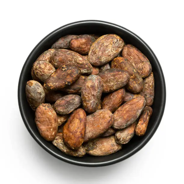 Roasted unpeeled cocoa beans in a black ceramic bowl isolated on white from above.