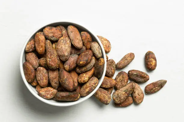 Roasted unpeeled cocoa beans in a white ceramic bowl next to a pile of unpeeled cocoa beans isolated on white painted wood  from above.