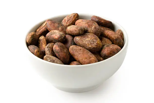 Roasted unpeeled cocoa beans in a white ceramic bowl isolated on white.