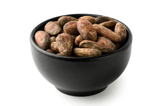 Roasted unpeeled cocoa beans in a black ceramic bowl isolated on white.