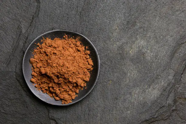 Cocoa powder in a black ceramic dish isolated on black slate from above. Space for text.