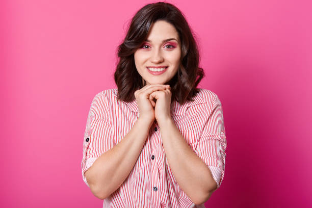 beautiful woman portrait, female with toothy charming smile, keeps hands under chin, looks directly at camera, wearing striped blouse, posing against pink studio wall. people emotions concept. - stripped shirt imagens e fotografias de stock