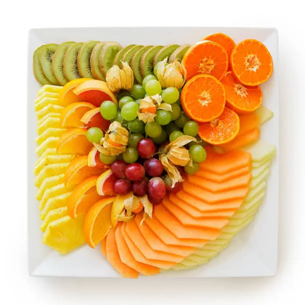 Mix of freshly cut tropical and citrus fruit arranged on white square ceramic platter isolated on white from above.