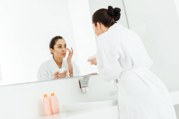 istock selective focus of attractive brunette woman applying face cream while looking at mirror 1153688534