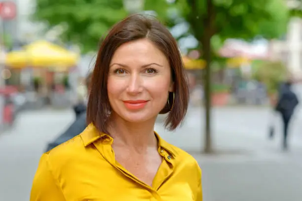 Attractive mature woman in colorful yellow shirt enjoying a day in town standing in a quiet urban square smiling at the camera