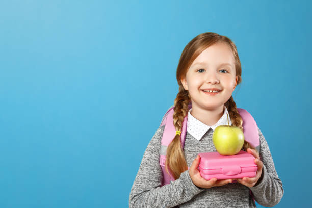 un retrato de una niña colegiala en un fondo azul. el niño tiene una mochila a sus espaldas y tiene una lonchera en sus manos. de vuelta a la escuela. el concepto de educación. - lunch box child education school fotografías e imágenes de stock