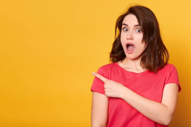 istock Studio shot of astonished black woman points aside with thumb, has thrilled expression, shocked to hear new track, wears casual black t shirt, isolated over yellow studio wall. Gosh, what news 1153696785