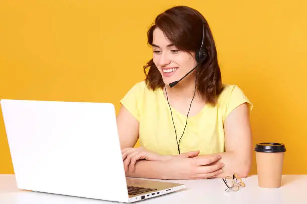 Smiling energetic female sitting at office desk with folded arms, having headset, looking at laptop screen, papercup of coffee and eyeglasses are laying on desk surface, wearing casual yellow t shirt.