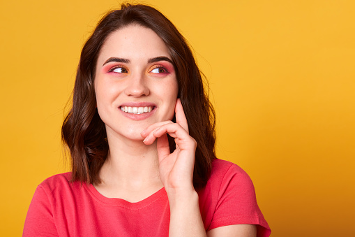 Close up studio portrait of pretty lovely charming lady looking away, keeps hand under chin, trying to understand or remember something, has bright make up, posing isolated over yellow background.