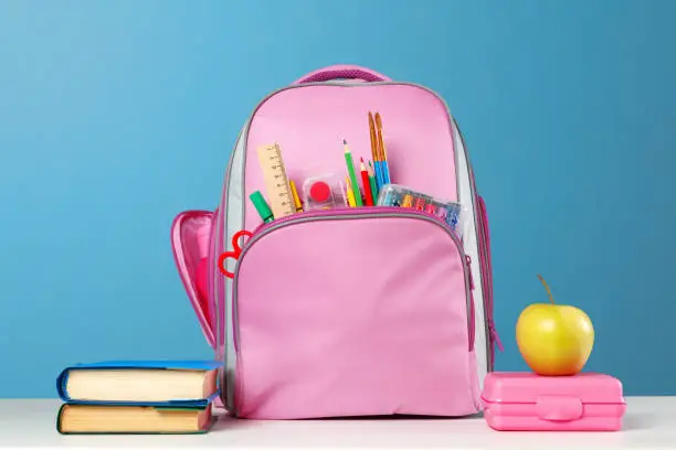 Student set. Pink backpack with stationery, a stack of books, a lunchbox, an apple on the table on a blue background. Back to school.