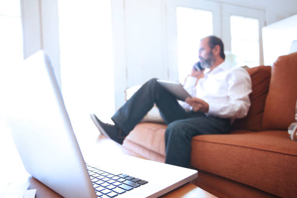 A Hispanic man in his late fifties sits on a couch in his home as he talks on his mobile device and holds a digital tablet that sits in his lap.  The man is blurred in the background with the focus being on a laptop computer that sits on the coffee table in front of him.  Light from outside filters through the French door  panes and fills the room with a soft glow.