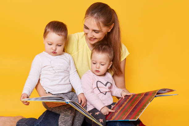 studio shot of happy family: mother and little twins girls sitting on floor, reading books and viewing bright interesting pictures, spending time with mommy, selebrating mother's day. children concept - love growth time of day cheerful imagens e fotografias de stock