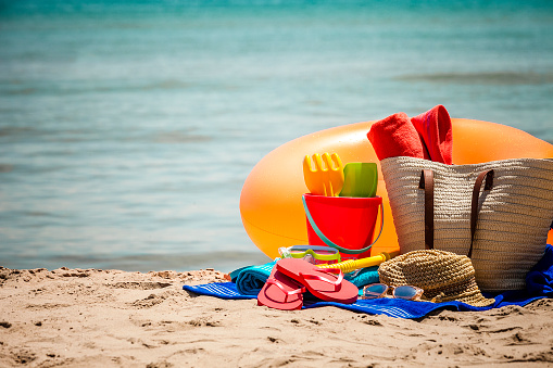 Front view of multi colored beach accessories shot on beach sand with defocused sea at background. Accessories included in the composition are beach towels, a pair of flip-flops, scuba mask with snorkel, beach bag, sun hat, sand bucket and sunglasses. The accessories are grouped at the right of the frame leaving useful copy space for text and/or logo. Beach, summer or vacations concept. DSRL outdoors photo taken with Canon EOS 5D Mk II and Canon EF 70-200mm f/2.8L IS II USM Telephoto Zoom Lens