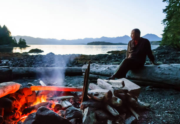 istock Man sitting on beach by a campfire in the wilderness, Broken Group Islands, Barkley Sound, Pacific Rim National Park, Canada. Middle aged mature people enjoying outdoor adventures. Selective focus on fire. 1153703426