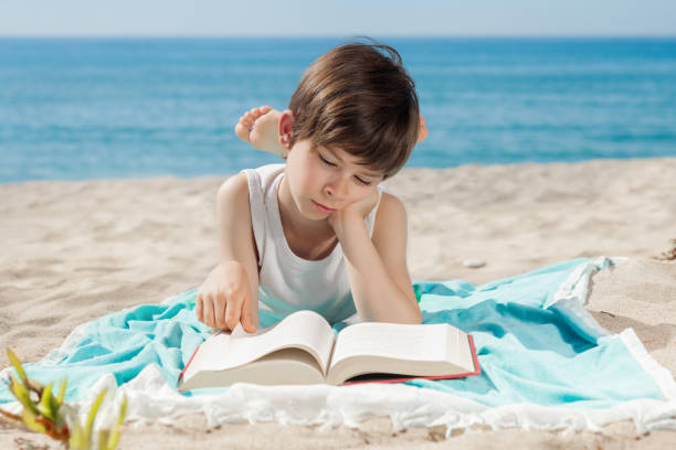 Cute boy reading a book on beach near sea at sunset