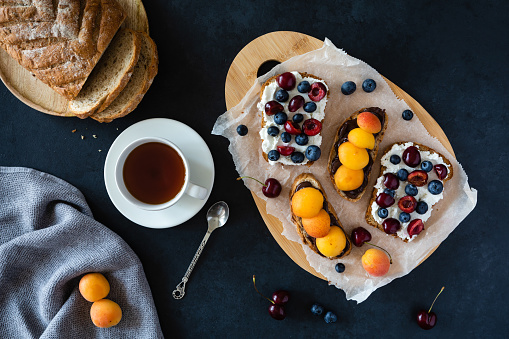Light summer breakfast or lunch with sandwiches of wholegrain bread with chocolate paste, ricotta cheese, apricots and sweet cherry on the background of the dark village table