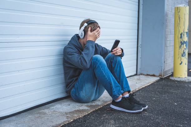 Sad teenager wearing headphones and listening to music. The image displays a sad teenage boy listening to music and viewing something upsetting on his cellphone. He is wearing a backpack, hoodie and headphones. sad music stock pictures, royalty-free photos & images