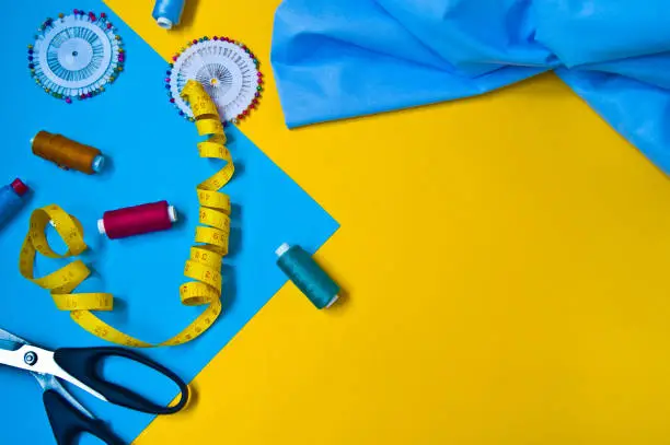 tools for creativity and needlework, sewing threads, scissors, sewing needles and centimeter tape on blue and yellow background, sewing accessories, craft, sewing clothes