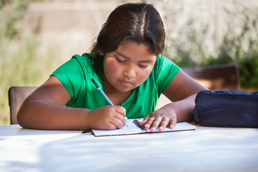 Focused young girl writting in her notebook, problem solving and studying outdoors in homeschool setting.