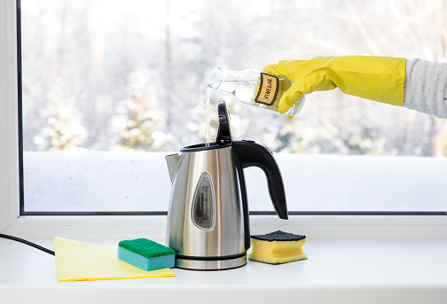 Woman pouring natural destilled acid white vinegar in electric kettle to remove boil away the limescale. Descaling a kettle, remove scale concept.