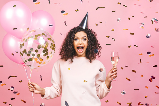 Celebration birthday concept - close up portrait of happy cheerful young beautiful african american woman with pink t-shirt with colorful party balloons and confetti, champagne. Isolated against pink studio Background.
