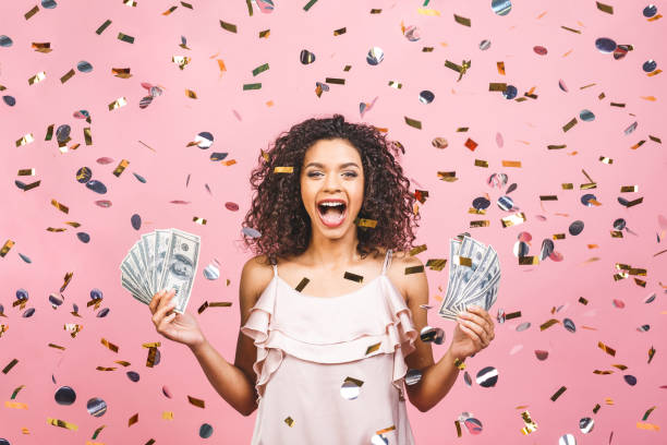 a menina afro-americana preta ganhou o dinheiro. mulher nova feliz que prende a moeda do dólar satisfeita isolada sobre o fundo cor-de-rosa com confetti. - jackpot - fotografias e filmes do acervo