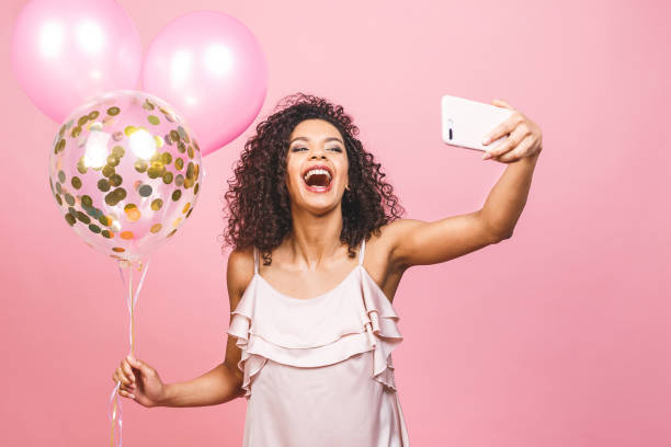 autoportret afro amerykańskiej pozytywnej dziewczyny w sukience mającej balony w ręku strzelające selfie na przednim aparacie odizolowanym na różowym tle. - party frock zdjęcia i obrazy z banku zdjęć