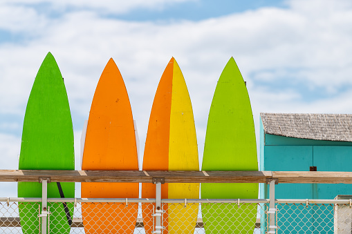 Stack row of multicolored colorful stand up surfing boards on railing fence with green orange and yellow color against blue sky lifeguard tower or rental shop