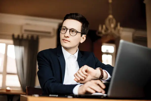 Photo of Portrait of a handsome young freelancer wearing glasses looking away while looking at his watch sitting in a coffee shop working at his laptop.