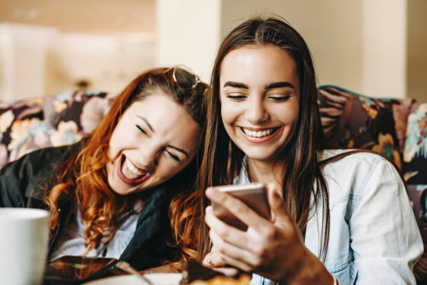 istock Beautiful young female holding a smartphone laughing while showing something to her girlfriends which is laughing leaning head on her shoulder. 1153731599