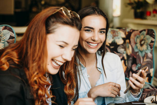 istock Lovely female with dark long hair looking at camera laughing while holding a smartphone while her girlfriends is looking down laughing in a coffee shop. 1153731606