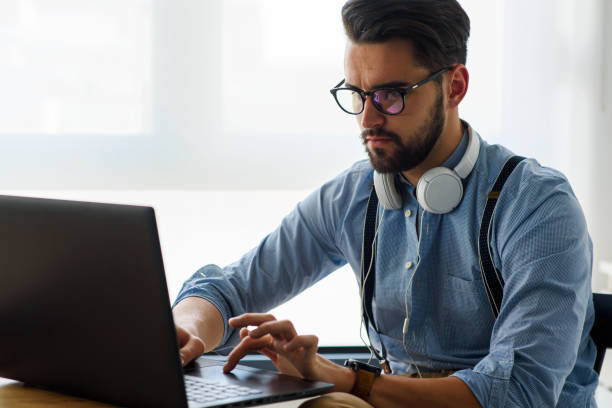 istock Young modern bearded man working with laptop computer indoors 1153733666