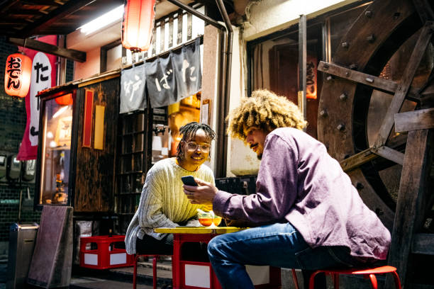 Hipster couple looking at smartphone outside restaurant Couple sitting outside Japanese Izakaya using mobile phone, social media, online messaging japanese girlfriends stock pictures, royalty-free photos & images