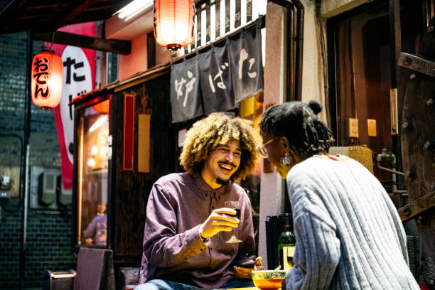 Attractive couple laughing with wine outside Japanese restaurant Young man with Afro hair laughing with friend and sitting outside Japanese Izakaya, fun, togetherness, nightlife tourist couple candid travel stock pictures, royalty-free photos & images