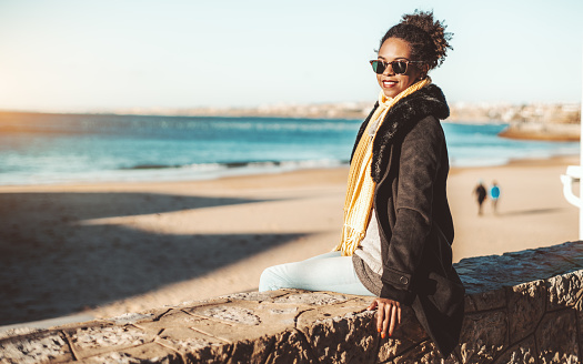 A happy young African-American female in sunglasses and coat is sitting on the stone fence wall on a sunny day with beach and water in the background and a copy space area on the left for your ad text