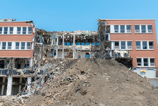 A partially demolished building. Much of the center of the building is missing, and there is a large pile of dirt in front. Lots of debris and hanging wires. Bright sunny day.