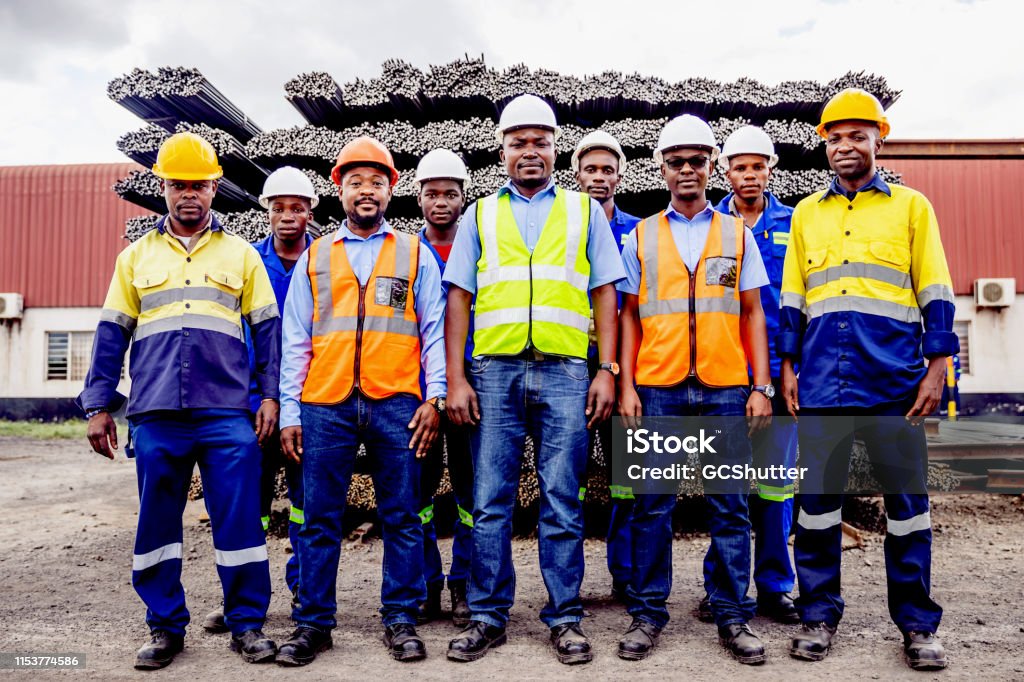 Group Portrait of Confident African Steel Factory Worker Team in Africa Africa, Industry, Business, Steel Factory, People - Portrait of a Factory Manual Worker, Supervisor, Safety Officer and Factory Manager Standing In front of the Steel Bar Storage Yard. Labor Union Stock Photo