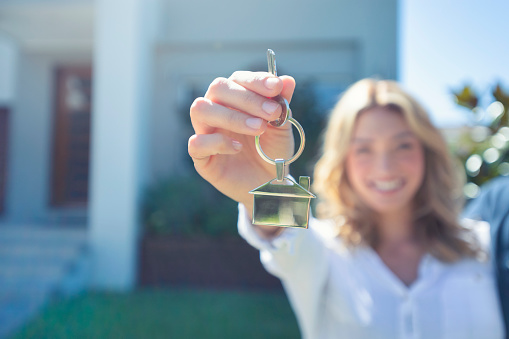 Young woman holding the key to her new house. She is smiling and happy. They keyring is shaped like a house. The house can be seen in the background.