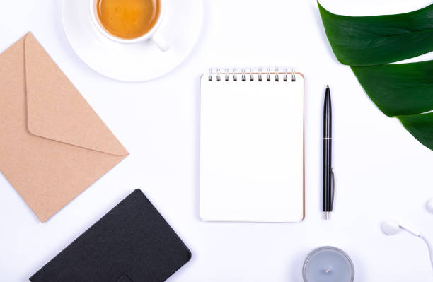 istock Composition of office workspace with tropical leaf plant Monstera, notebook, candle, pen, craft envelope, earphones and cup of coffee on white background. Business, educational
 or home office concept. 1153825431
