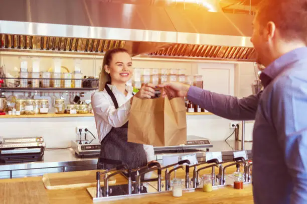 Cheerful waitress wearing apron serving customer at counter in restaurant - Small business and service concept with young woman owner giving eco friendly paper bag with take away food to online client
