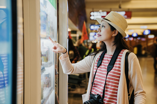 asian travel woman photographer using modern vending machine in airport while waiting for plane flight in departure gate. young girl right hand finger pointing choosing snack drinks to buy.