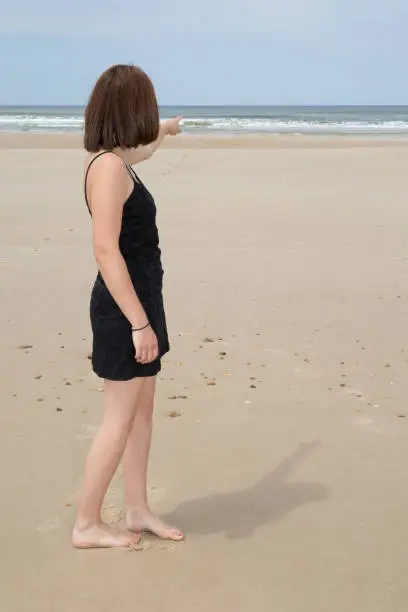 Young brunette woman in summer blackdress standing on beach and looking to the sea. Caucasian girl relaxing and enjoying peace on vacation.