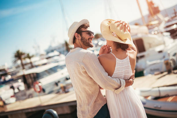 feliz pareja joven caminando por el puerto de un resort turístico en el mar con veleros en el fondo - bulevar fotos fotografías e imágenes de stock