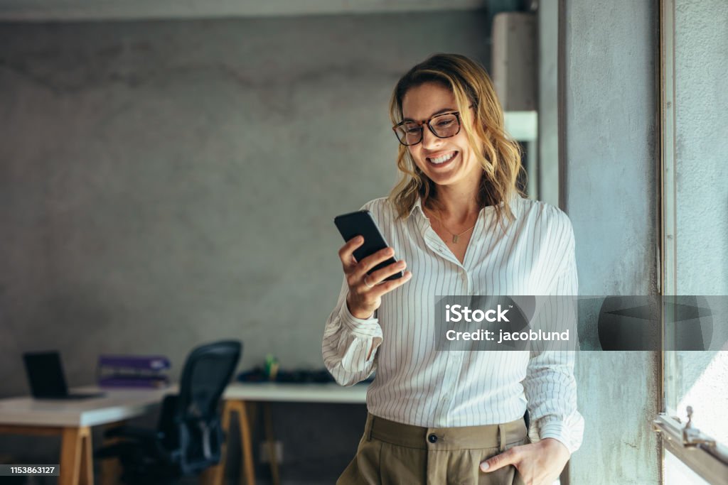 Smiling businesswoman using phone in office Smiling businesswoman using phone in office. Small business entrepreneur looking at her mobile phone and smiling. Women Stock Photo
